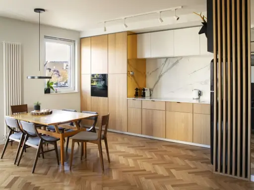 Drewniane lamele w kuchni przy stole tworzą przytulną atmosferę. Jasne fronty szafek i marmurowy blat dodają elegancji.