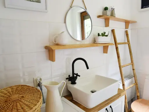 Urocza, mała łazienka w stylu retro z białymi kafelkami, drewnianymi półkami, okrągłym lustrem i bambusową drabiną.