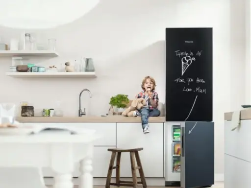 Chłopiec z pluszowym psem siedzi na blacie w kuchni, jedząc lody. Obok lodówka z tablicą, na której mama napisała "There's an ice cream for you here". Inspiracje na tablicę w kuchni.