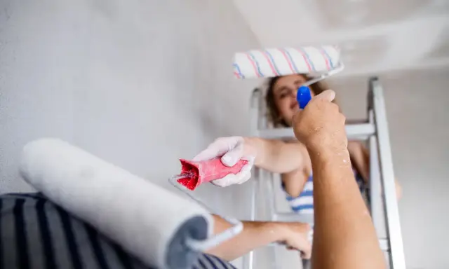 Dwie osoby malują sufit. Jedna stoi na drabinie, druga podaje jej wałek. Trwa nakładanie akrylu przy suficie.