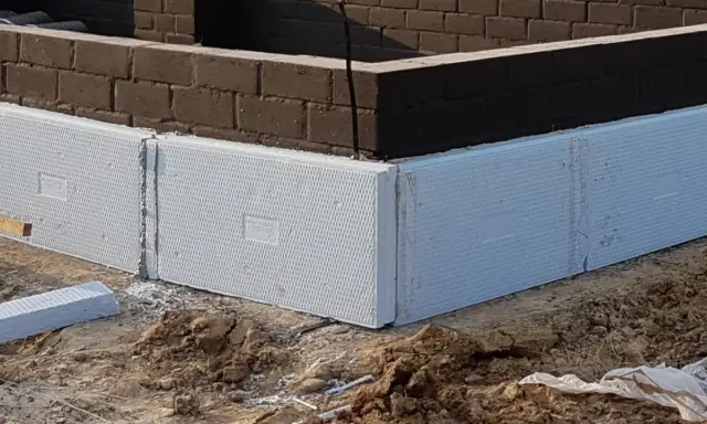 Białe, ekstrudowane bloki styropianowe tworzą fundament domu, otaczając ciemną ceglaną ścianę.