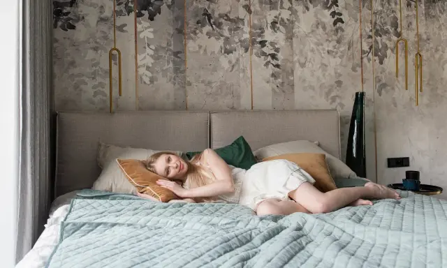 Młoda kobieta odpoczywa na łóżku. W tle widać stylową tapetę z motywem liści, która sprawia, że tanie tapety do sypialni wyglądają luksusowo.