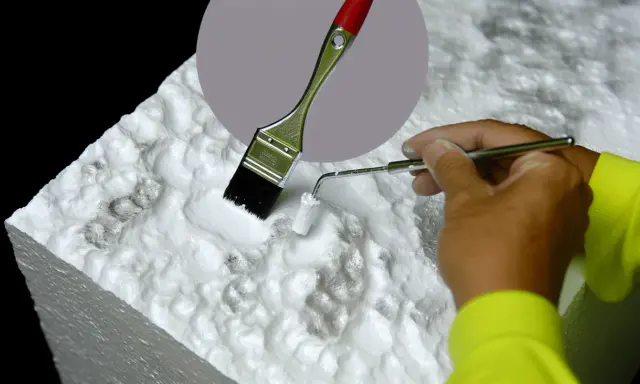 Ręka w żółtej bluzie maluje białą farbą styropian za pomocą pędzla i małego wałka. Dowiedz się, czym pomalować styropian.