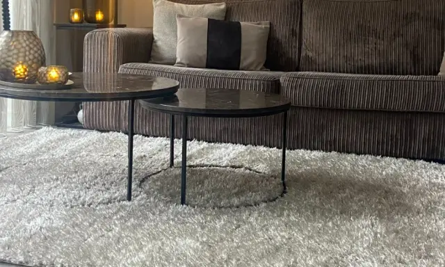 Miękki dywan w kolorze greige otula salon. Na nim dwa stoliki kawowe z marmurowymi blatami i aksamitna kanapa.