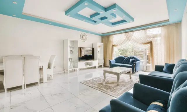 Nowoczesny salon z błękitnym, geometrycznym, dekoracyjnym malowidłem na suficie. Wygodne, granatowe sofy i białe meble tworzą elegancką przestrzeń.