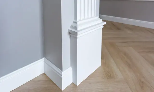 Biały pilaster z żłobieniami i elegancką bazą, idealny do wnętrzarskiego bloga. Drewniana podłoga w jodełkę i szara ściana tworzą stylowe tło.