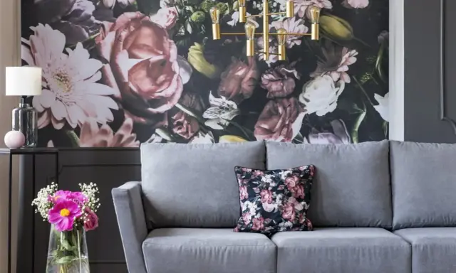 Szara sofa z poduszką w kwiaty, na tle bogatej fototapety. Idealna, by stworzyć przytulną atmosferę.