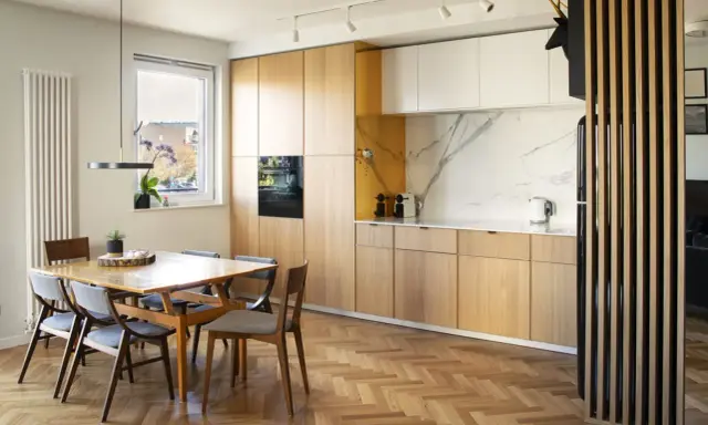 Drewniane lamele w kuchni przy stole tworzą przytulną atmosferę. Jasne fronty szafek i marmurowy blat dodają elegancji.