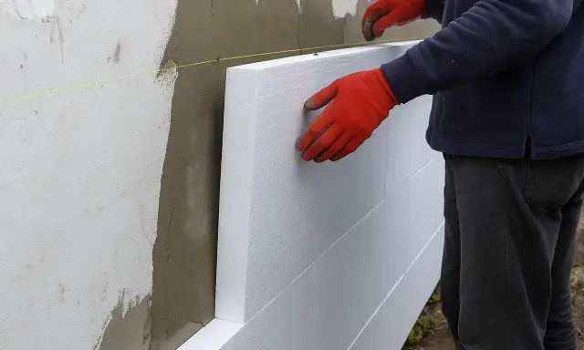 Pracownik w czerwonych rękawicach układa płyty styropianowe, rozpoczynając naprawę elewacji styropianowej.
