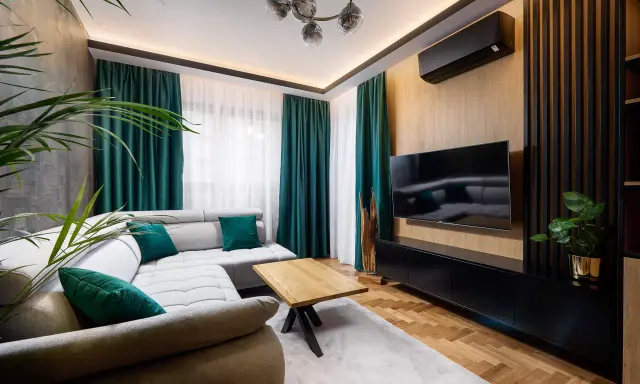 Nowoczesny salon z szarą kanapą i zielonymi poduszkami. Po prawej stronie ściana ozdobiona pionowymi lamelami w narożniku, na niej telewizor.