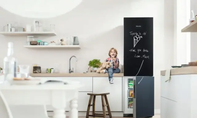 Chłopiec z pluszowym psem siedzi na blacie w kuchni, jedząc lody. Obok lodówka z tablicą, na której mama napisała "There's an ice cream for you here". Inspiracje na tablicę w kuchni.