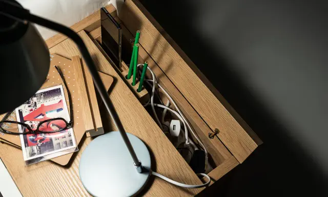 Drewniana szafka nocna z przegródkami, gdzie można schować wystające kable ze ściany. Na blacie lampka, okulary i notatnik.