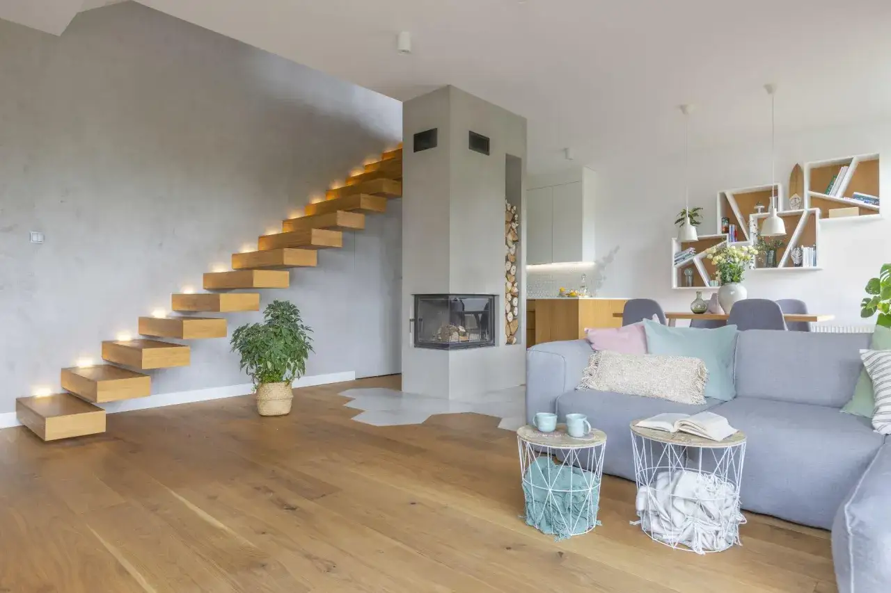 Nowoczesny salon z betonową ścianą, pływającymi schodami i kominkiem. Beton w salonie inspiracje: szara ściana, drewniane stopnie, szara kanapa.
