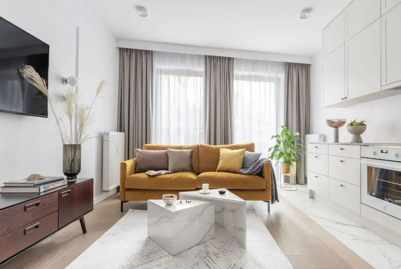Sofa w kolorze musztardowym, otoczona szarymi zasłonami i białymi meblami kuchennymi.