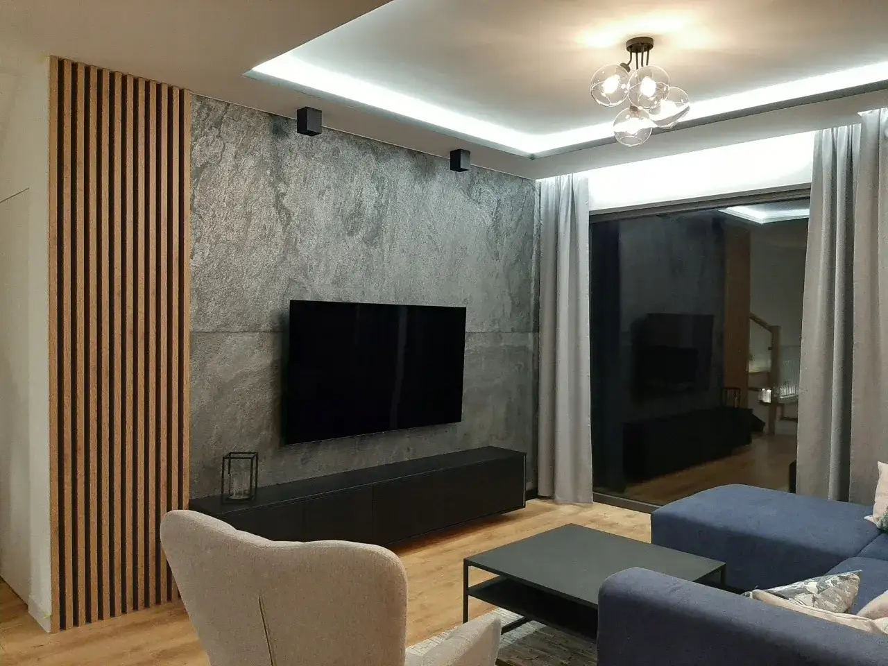 Nowoczesny salon z dominującą szarą ścianą, ozdobioną drewnianymi lamelami i kamieniem. Centralnie umieszczony telewizor i wygodna kanapa zapraszają do relaksu.