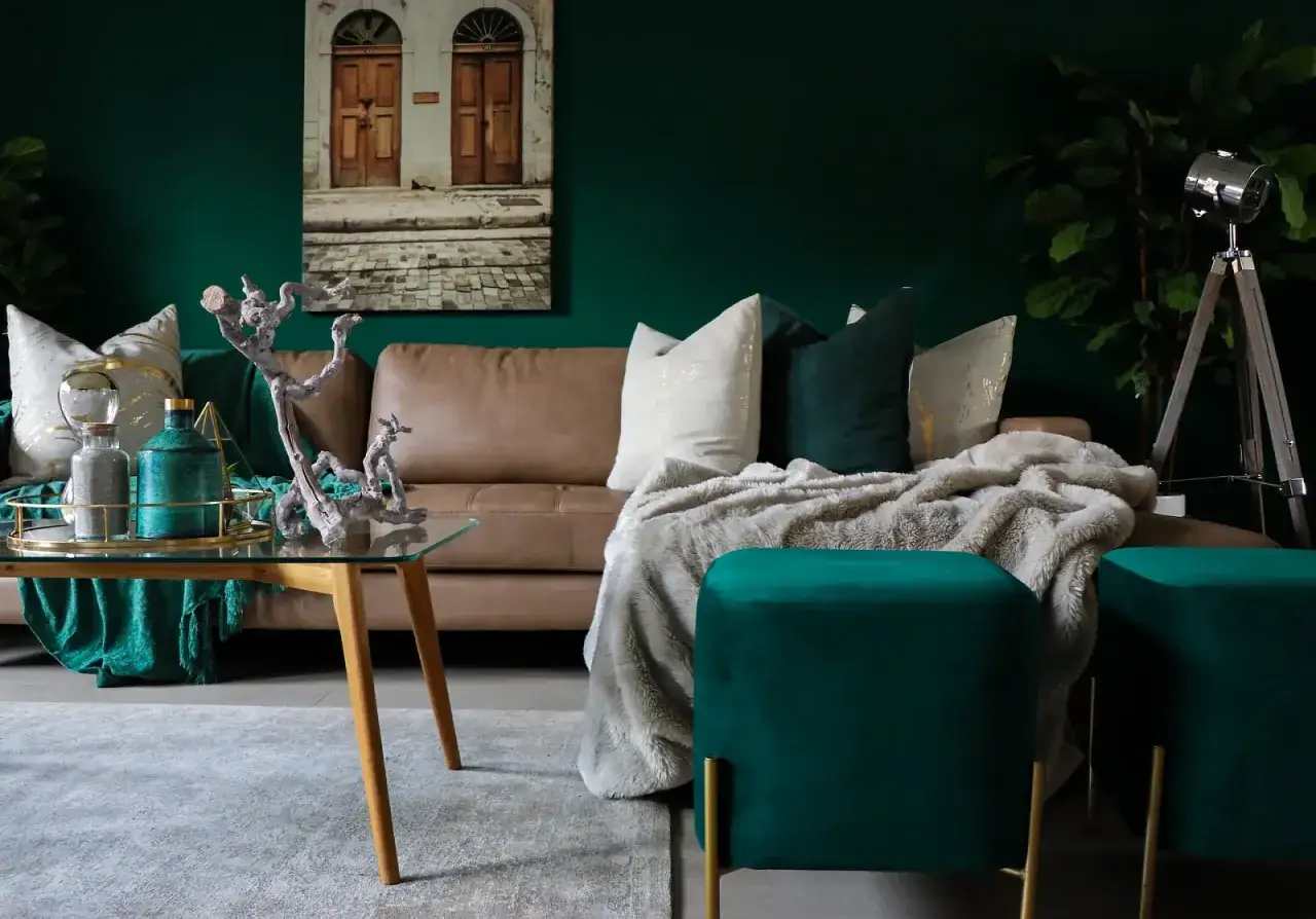 Elegancki salon z sofą, puszystym kocem i pufami. Wzrok przyciągają kolor ścian butelkowa zieleń oraz dekoracje.