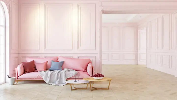 Eleganckie wnętrze w pastelowym różu z ozdobną sztukaterią na ścianach. Miękka kanapa, stoliki kawowe i drewniana podłoga tworzą przytulną atmosferę.