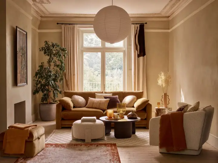 Przytulny salon z kanapą w kolorze karmelowym, ozdobioną poduszkami. Obok fotel, puf i stolik kawowy.